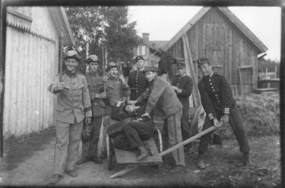 7 uniformsklädda män stående runt kärra och 1 man liggande på den.
Nyckelord: Militär Kärra Uthus