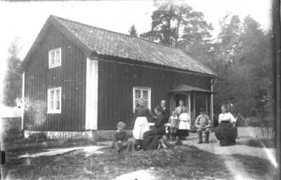 Ett antal personer stående eller sittande framför byggnad.
Nyckelord: Parkbänk