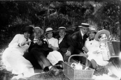 4 män och 3 kvinnor sittande på backen, en med dragspel i knät.
Nyckelord: Dragspel Picnic