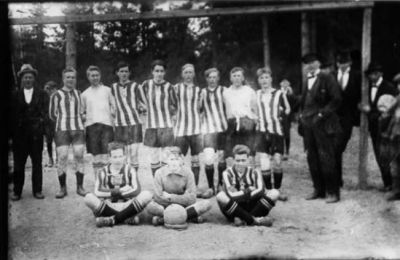 11 fotbollsklädda män stående eller sittande med ytterligare män stående bredvid.
Nyckelord: Dräkt Fotboll