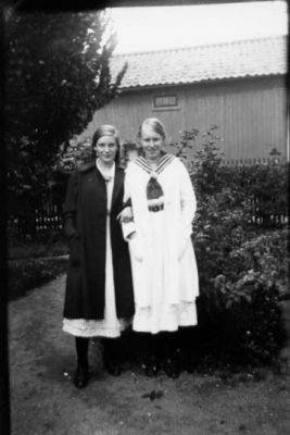 2 flickor stående i trädgård.
