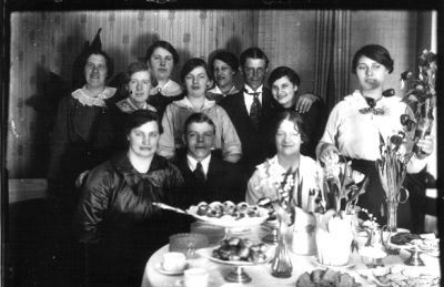 Man och 2 kvinnor sittande, man och 7 kvinnor stående vid kaffebord i rum.
Nyckelord: Kaffebord Blommor