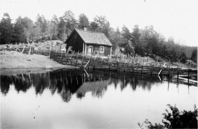 Byggnad bakom gärdesgård och vatten.
Nyckelord: Vatten Gärdesgård