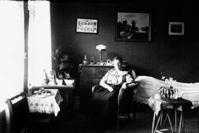 Kvinna sittande i korgstol i rum.
Nyckelord: Lampa Tavla Sängöverkast