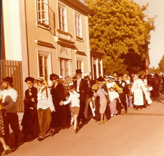 Festklädda människor i kö utmed byggnad
Nyckelord: Dräkt