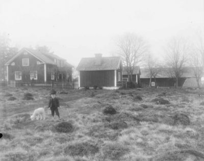 Pojke och lamm på gärde framför byggnader
Nyckelord: Dräkt Lamm