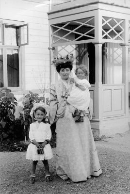 Kvinna och 2 barn på gårdsplan framför byggnad
Nyckelord: Dräkt Hatt